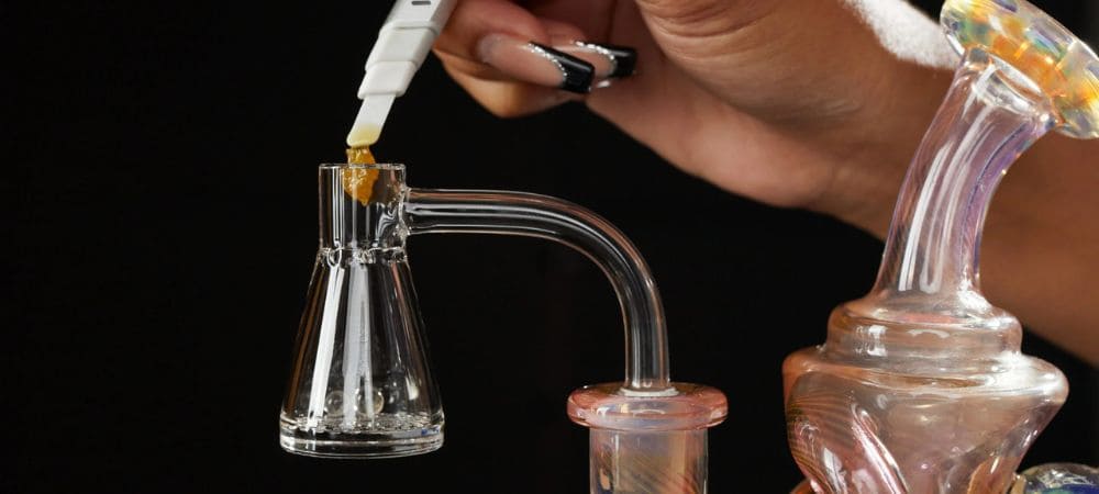 Primo piano di una mano che posiziona il concentrato su un banger in vetro trasparente montato su un dab rig, con sfondo scuro.
